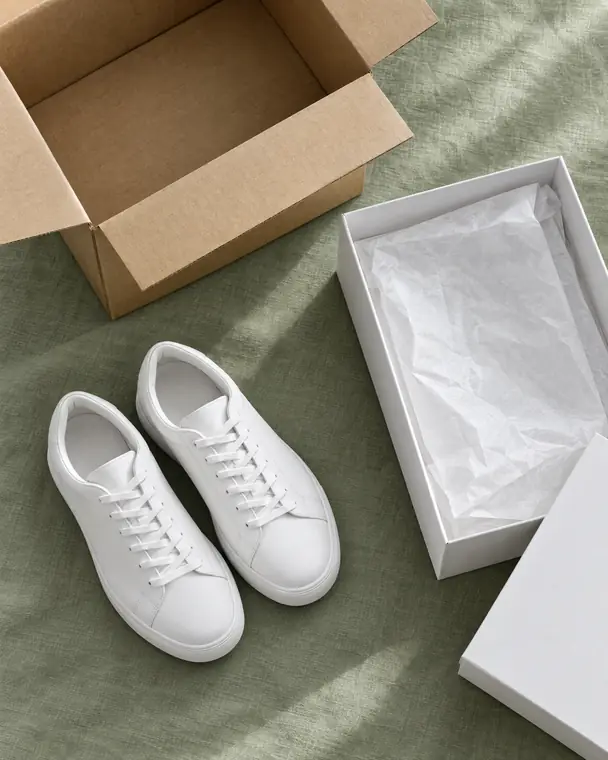 A pair of white sneakers next to a shoe box and a shipping box.